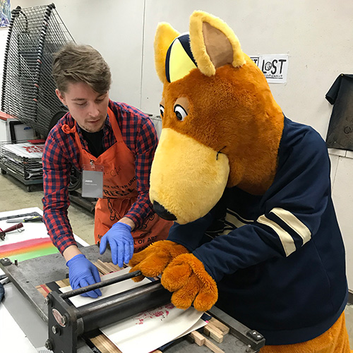 A Myers School of Art student wearing a printmaking apron and gloves demonstrates printmaking techniques to Zippy, The University of Akron mascot, as they operate a printing press together in a campus art studio. Printing equipment and drying racks are visible in the background.